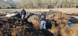 Two workers in branded hoodies dig and prepare piping in a dirt trench at a residential construction site with a house and woods in the background.